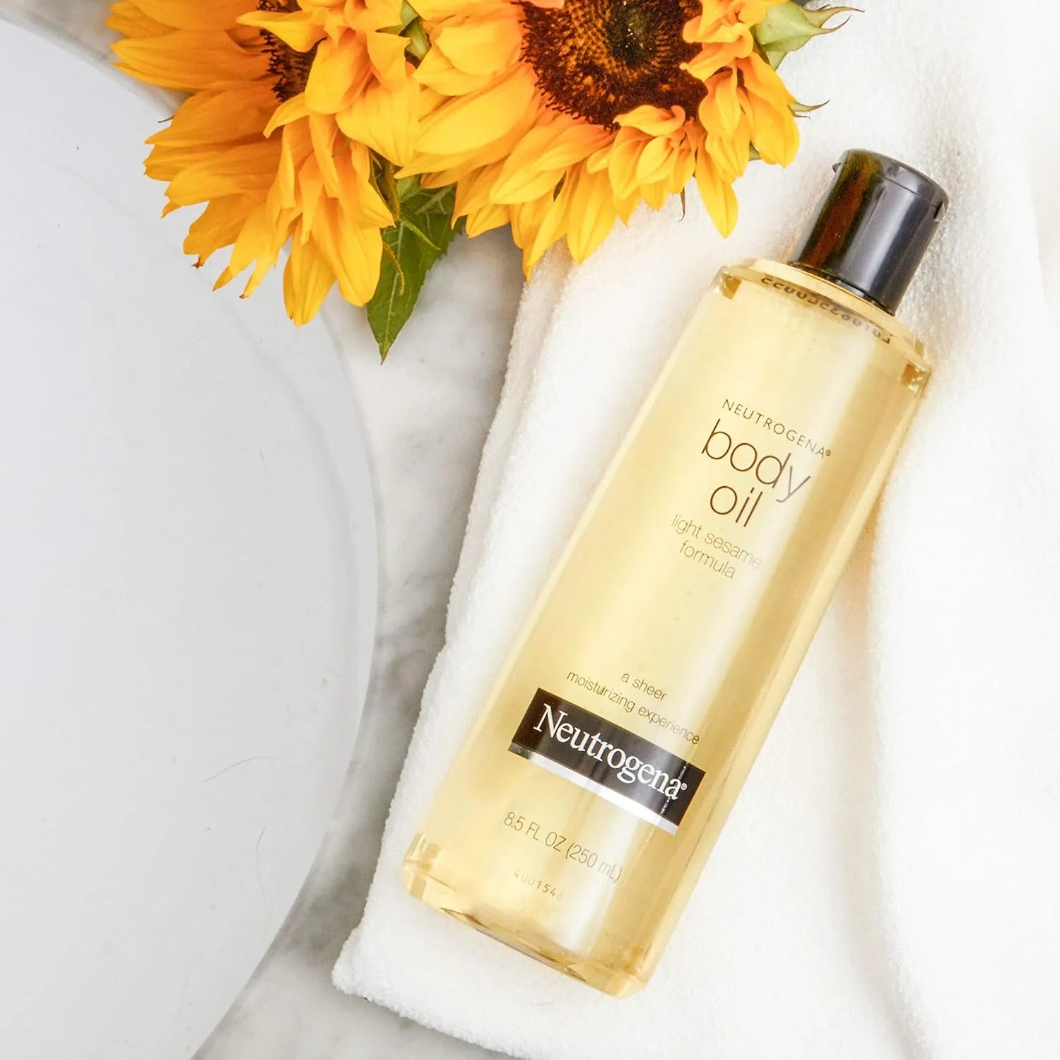 Neutrogena Body Oil Light Sesame Formula 16 fl oz bottle placed on white towel beside sunflowers.